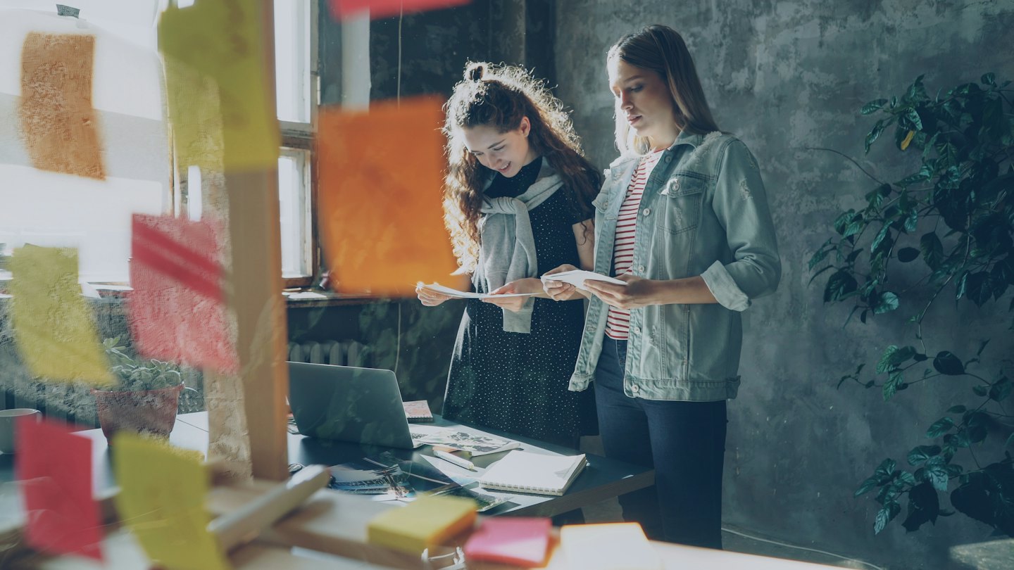 Dos mujeres revisan documentos y un portátil en un escritorio de oficina.